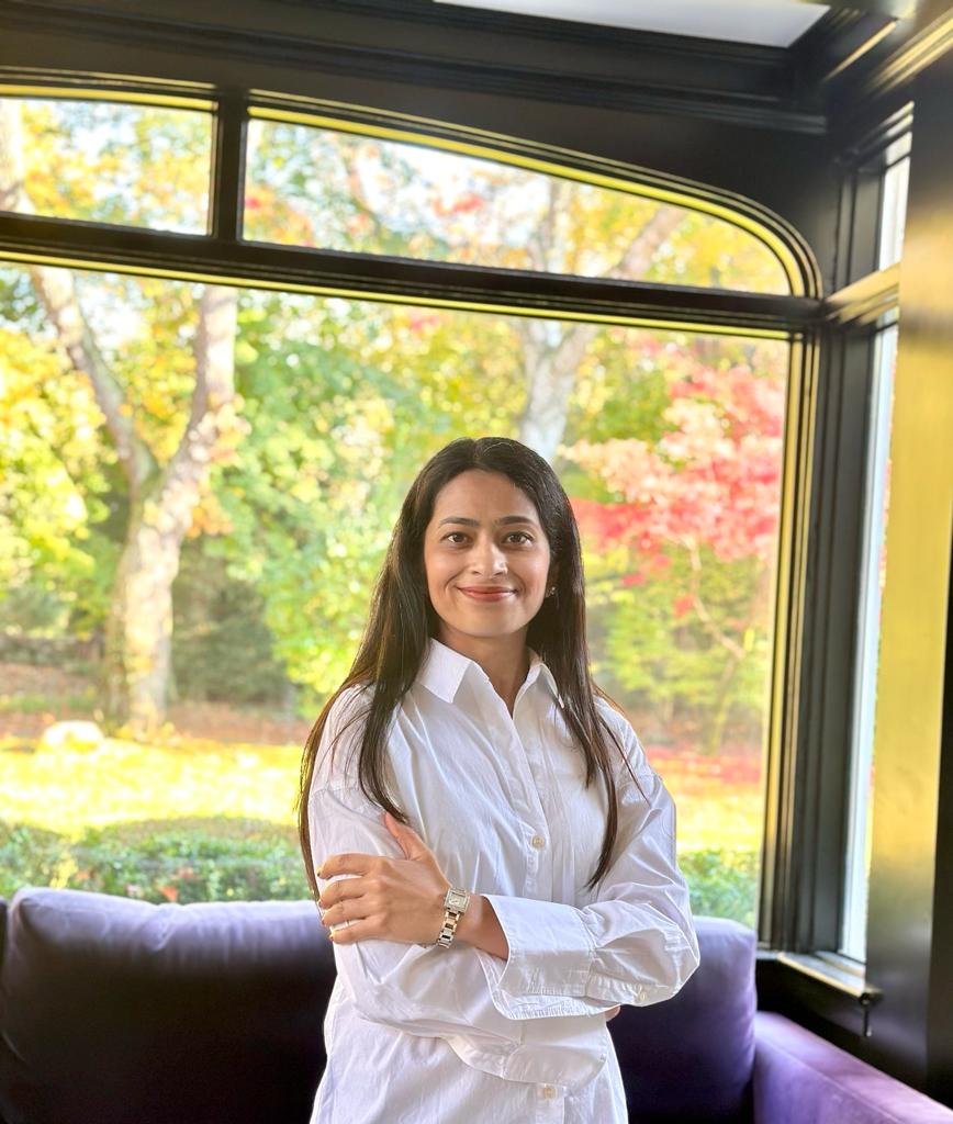 Pragya Jindal smiling in front of a sunlit window with autumn trees behind her